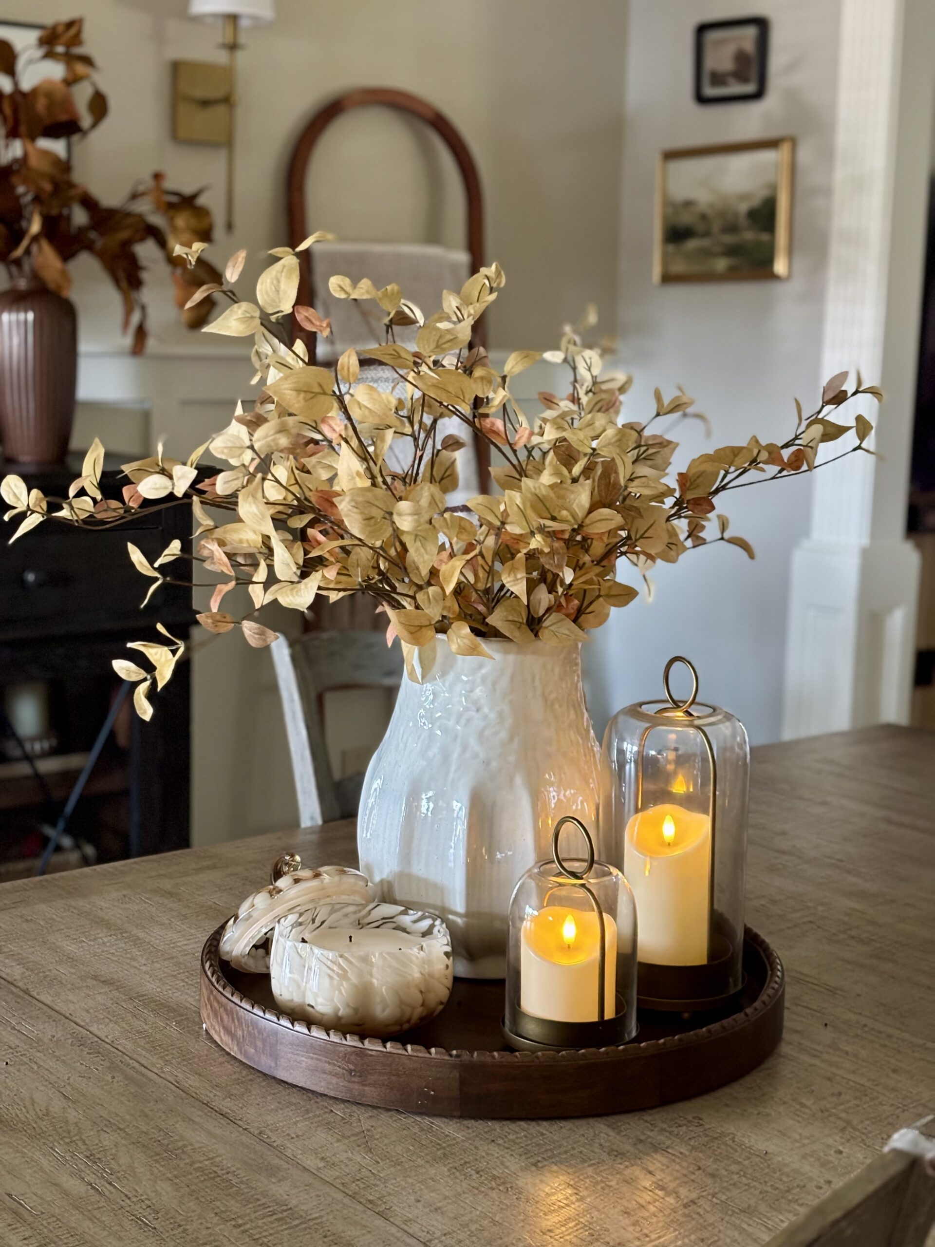 Fall dining room centerpiece with vase, seasonal stems, glass cloche candle holders, and pumpkin candle on a tray